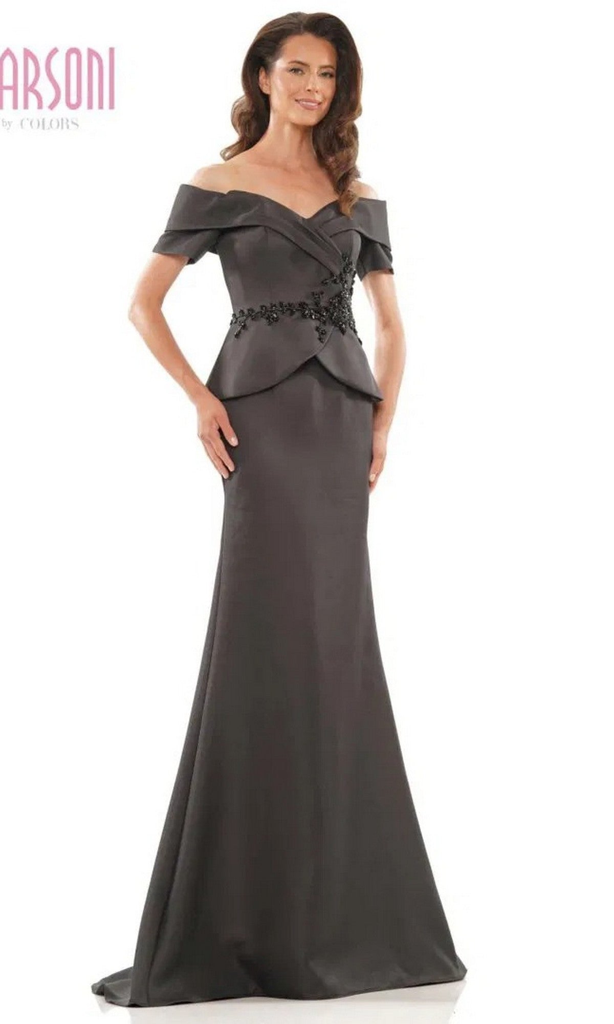 A woman wears the Marsoni MV1144, a long black formal dress featuring an off-the-shoulder neckline, fitted bodice, beaded peplum detail, and elegant embellishments at the waist—perfect for evening events.